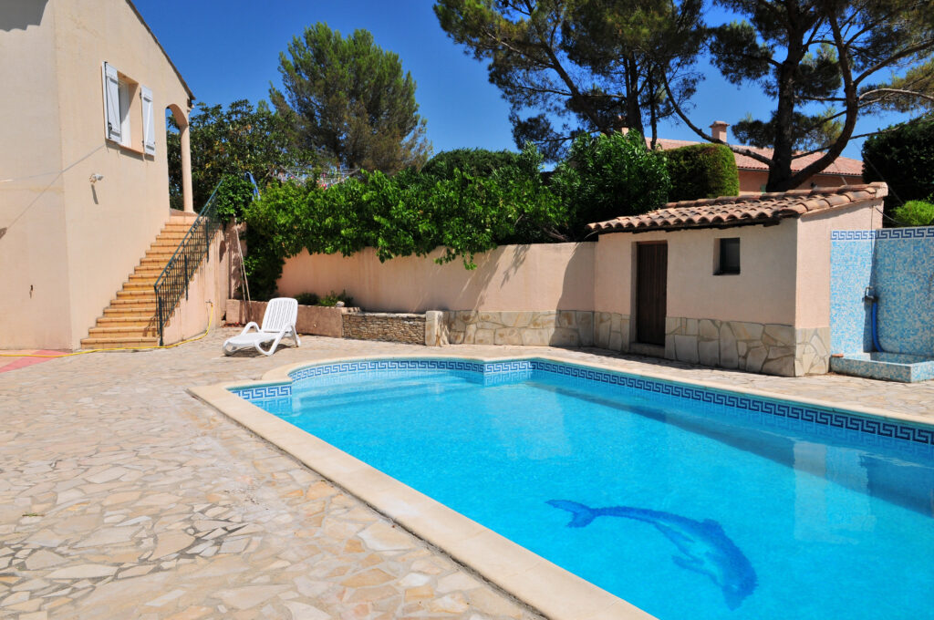 Grande piscine bleue avec mosaïque dauphin devant une maison méditerranéenne ensoleillée et son jardin. Parfait pour l'été.