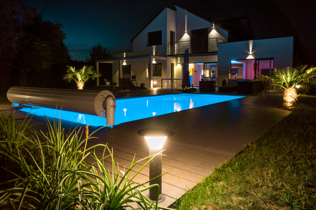 Maison moderne de nuit avec piscine bleue lumineuse, terrasse bois et éclairage extérieur. Ambiance chic et relaxante.