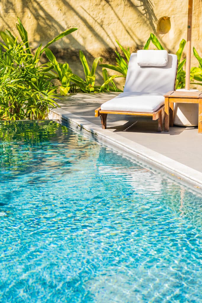 Piscine bleue scintillante, transat blanc et table en bois sous le soleil. Végétation luxuriante près d'un mur texturé, ambiance détente tropicale.
